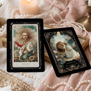 Puede incluir: Dos cartas de tarot con bordes negros se muestran sobre una superficie suave y texturizada. Una carta presenta un sol con rostro, la otra muestra a una mujer con un león. Las cartas están rodeadas de velas, perlas y flores secas.