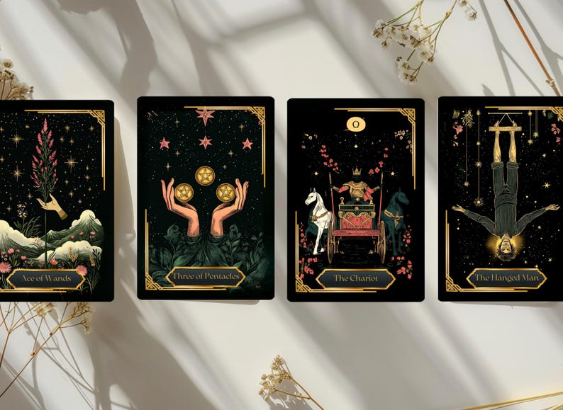 May include: Four tarot cards with black backgrounds and gold accents. The cards feature illustrations of the Ace of Wands, Three of Pentacles, The Chariot, and The Hanged Man. Each card has a unique design with celestial and mystical elements.