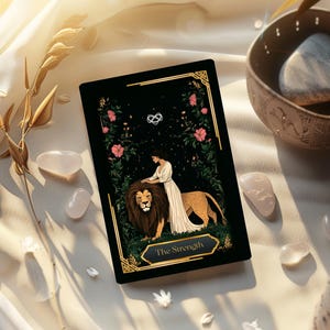May include: A tarot card with a black background featuring a woman and a lion, labeled "The Strength." Pink flowers and gold accents frame the image. The card rests on a white cloth with quartz crystals and a decorative bowl.