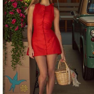 Pu&ograve; includere: Un vestito rosso senza maniche con bottoni. L'abito &egrave; abbinato a un cestino di vimini. Lo sfondo include un veicolo vintage verde e una composizione floreale. L'abito &egrave; in stile mini.