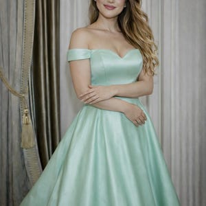 Puede incluir: Un vestido de fiesta verde menta con hombros descubiertos y escote coraz&oacute;n. El vestido tiene una falda amplia y est&aacute; hecho de una tela lisa, similar al sat&eacute;n. La modelo lleva el vestido delante de un fondo neutro.