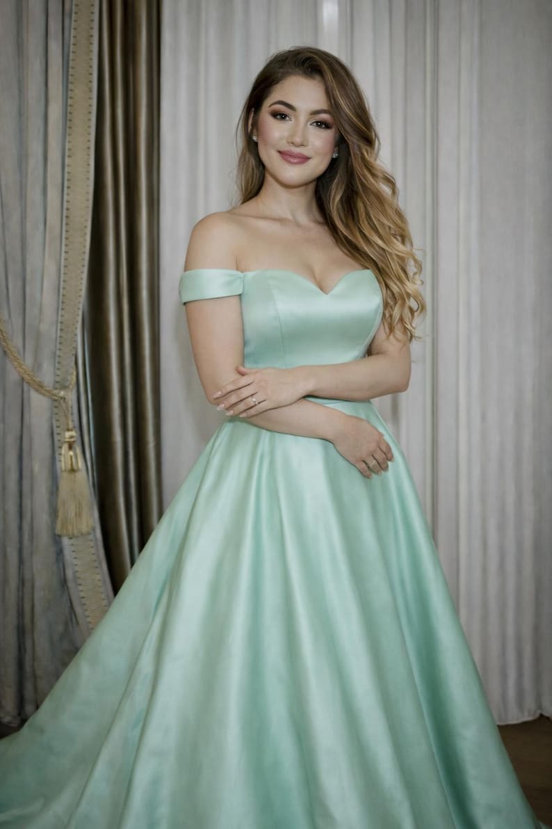 Puede incluir: Un vestido de fiesta verde menta con hombros descubiertos y escote coraz&oacute;n. El vestido tiene una falda amplia y est&aacute; hecho de una tela lisa, similar al sat&eacute;n. La modelo lleva el vestido delante de un fondo neutro.