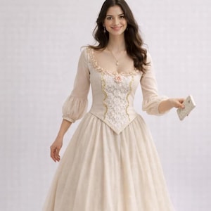 May include: A cream-colored, long-sleeved dress with a square neckline and a full skirt. The bodice features intricate lace and gold trim. The sleeves are sheer with a puffed design. A small pink flower is at the center of the bodice.