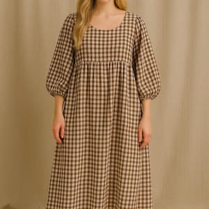 Peut inclure: Robe à carreaux marron et blanc avec un col rond et des manches trois-quarts. La robe a une taille froncée et une jupe midi. Le modèle porte la robe. L'arrière-plan est de couleur neutre.