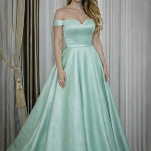 Puede incluir: Un vestido verde menta con hombros descubiertos, escote coraz&oacute;n y falda amplia. El vestido tiene un corpi&ntilde;o ajustado y una falda fluida, creando una silueta elegante. La tela tiene un brillo sutil y el vestido es largo.