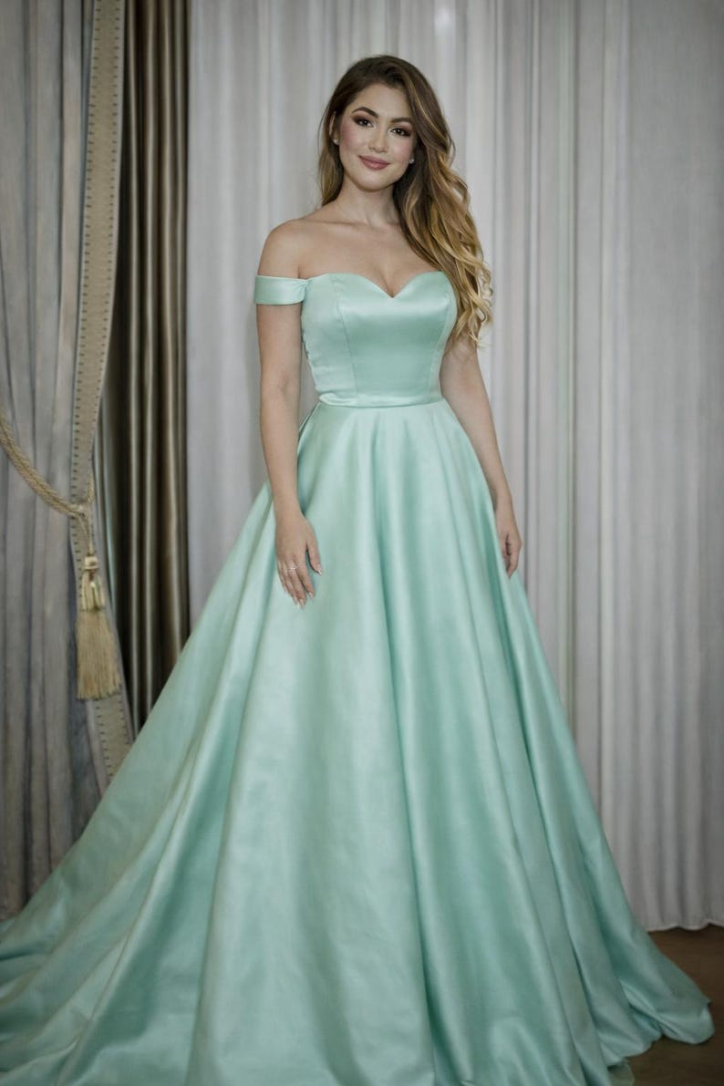 Puede incluir: Un vestido verde menta con hombros descubiertos, escote coraz&oacute;n y falda amplia. El vestido tiene un corpi&ntilde;o ajustado y una falda fluida, creando una silueta elegante. La tela tiene un brillo sutil y el vestido es largo.