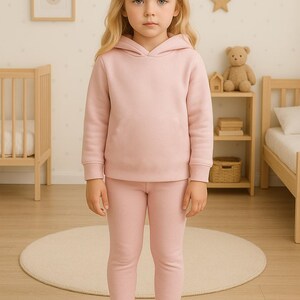 May include: A young child wearing a matching pink hoodie and leggings set. The outfit is a solid color, and the child is standing on a light-colored rug. The background includes a wooden bed frame, a small shelf with a teddy bear, and star-shaped wall decorations.
