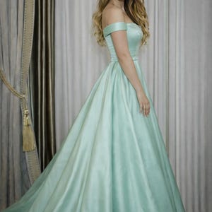 Puede incluir: Un vestido verde menta con hombros descubiertos y falda fluida. El vestido tiene un corpi&ntilde;o ajustado y una falda larga hasta el suelo. La tela tiene un brillo sutil. La modelo lleva el vestido delante de un fondo neutro.