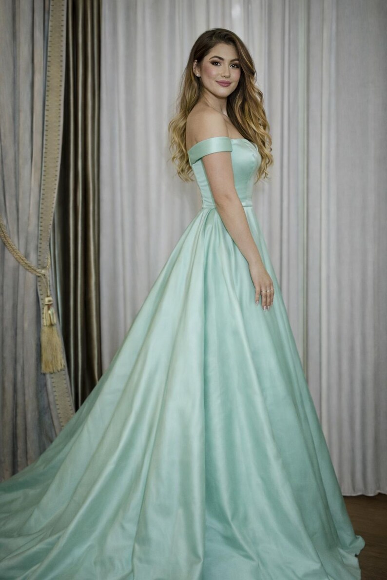 Puede incluir: Un vestido verde menta con hombros descubiertos y falda fluida. El vestido tiene un corpi&ntilde;o ajustado y una falda larga hasta el suelo. La tela tiene un brillo sutil. La modelo lleva el vestido delante de un fondo neutro.