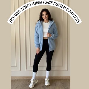 May include: A light blue hooded teddy sweatshirt with a zipper, worn over a white top and black leggings. The text "HOODED TEDDY SWEATSHIRT SEWING PATTERN" is arched above the model.