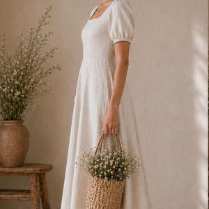 May include: A cream-coloured midi dress with short puff sleeves, a square neckline, and a fitted bodice that flows into a full skirt. The dress is made of a textured fabric and is paired with beige flat shoes and a woven straw basket filled with flowers.