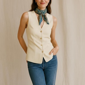 May include: A cream-colored vest with a button-down front is paired with blue jeans. A patterned teal scarf is worn around the neck. The outfit is simple and stylish.