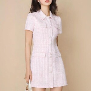May include: A pink tweed dress with a collared neckline, short sleeves, and a button-up front. The dress has a fitted silhouette and a slightly flared skirt.