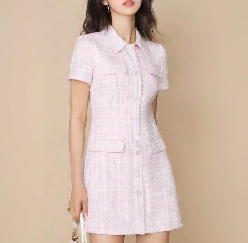 May include: A pink tweed dress with a collared neckline, short sleeves, and a button-up front. The dress has a fitted silhouette and a slightly flared skirt.