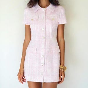 May include: A pink tweed dress with a button-up front and short sleeves. The dress has two pockets on the front and a white collar.