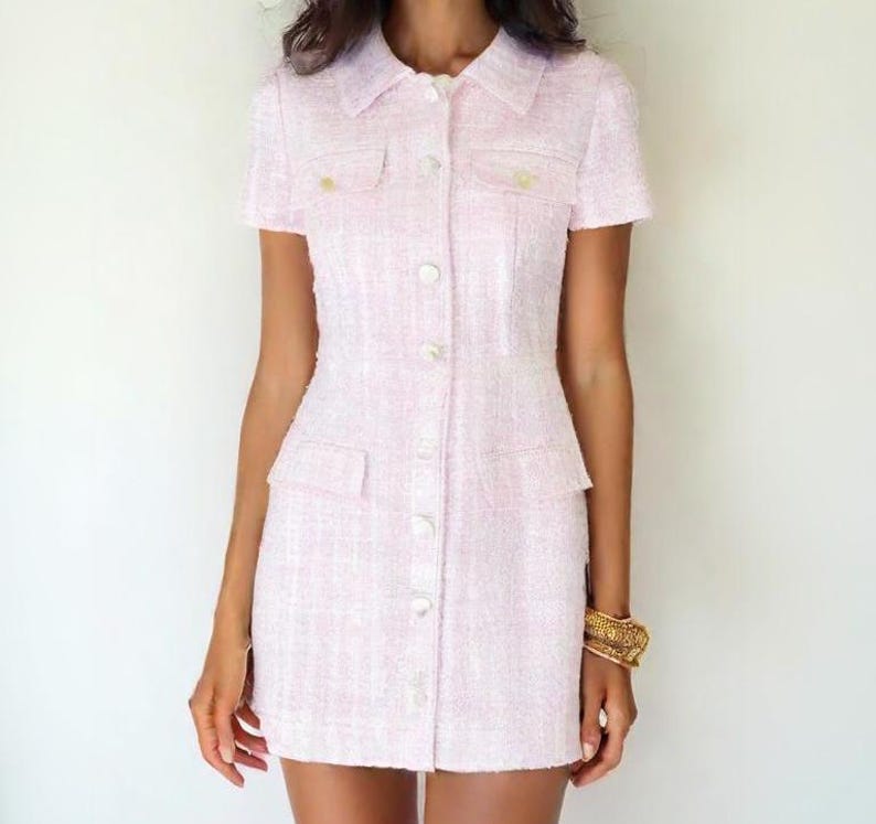 May include: A pink tweed dress with a button-up front and short sleeves. The dress has two pockets on the front and a white collar.