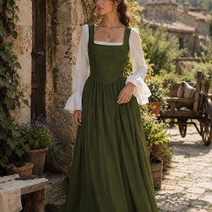 May include: A floor-length olive green dress featuring a fitted bodice with a square neckline and a full skirt, layered over a white long-sleeved blouse with flared cuffs. The dress has a structured waistline and a simple, solid-colored design.