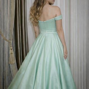 Puede incluir: Un vestido verde menta con hombros descubiertos y falda amplia. El vestido presenta un corpi&ntilde;o ajustado y una falda fluida, hecha de una tela lisa, similar al sat&eacute;n. La modelo se ve de espaldas, con pelo largo y rubio ondulado.