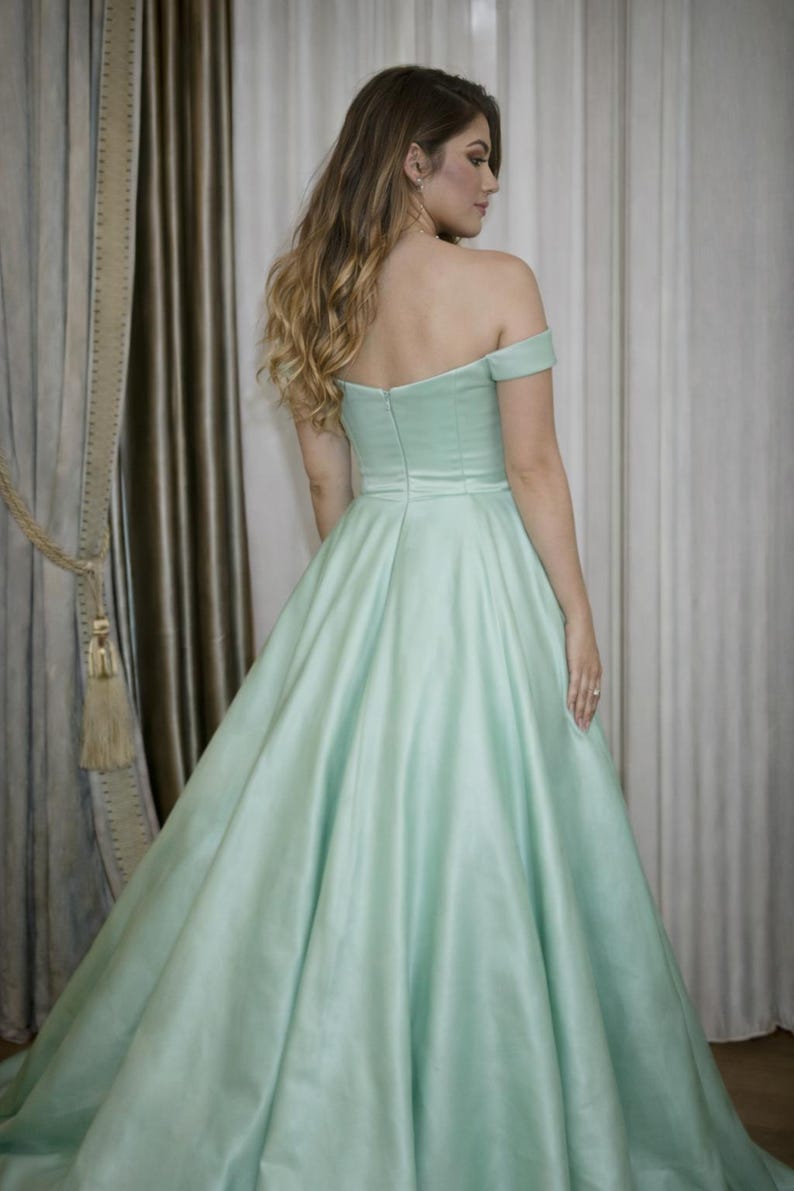 Puede incluir: Un vestido verde menta con hombros descubiertos y falda amplia. El vestido presenta un corpi&ntilde;o ajustado y una falda fluida, hecha de una tela lisa, similar al sat&eacute;n. La modelo se ve de espaldas, con pelo largo y rubio ondulado.