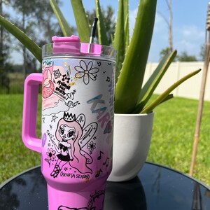 Op de afbeelding: Een roze roestvrijstalen beker met een zwarte rietje en een roze deksel. De beker heeft een witte achtergrond met een cartoon fee ontwerp en de tekst "Bichota Season".