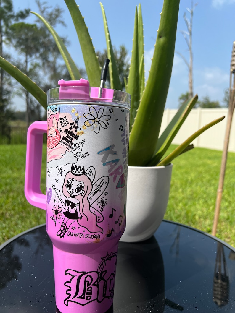 Op de afbeelding: Een roze roestvrijstalen beker met een zwarte rietje en een roze deksel. De beker heeft een witte achtergrond met een cartoon fee ontwerp en de tekst "Bichota Season".