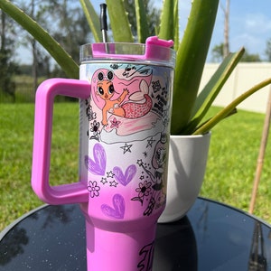 Op de afbeelding: Een roze roestvrijstalen beker met een doorzichtig deksel en een zwarte rietje. De beker heeft een kleurrijk ontwerp met twee cartoonmeisjes, harten, sterren en bloemen. De beker heeft een roze handvat.