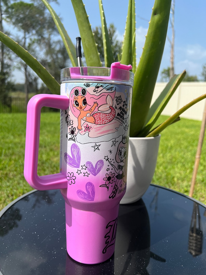 Op de afbeelding: Een roze roestvrijstalen beker met een doorzichtig deksel en een zwarte rietje. De beker heeft een kleurrijk ontwerp met twee cartoonmeisjes, harten, sterren en bloemen. De beker heeft een roze handvat.