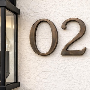 Puede incluir: Número de casa "02" de color bronce montado en una pared blanca texturizada. Los números están en una fuente clásica y están colocados a la derecha de una farola negra.