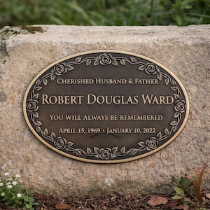 May include: A bronze memorial plaque with an oval shape, set into a stone. The plaque is inscribed with the words "CHERISHED HUSBAND & FATHER" and "ROBERT DOUGLAS WARD". It also includes the dates "APRIL 15, 1969 - JANUARY 10, 2022".