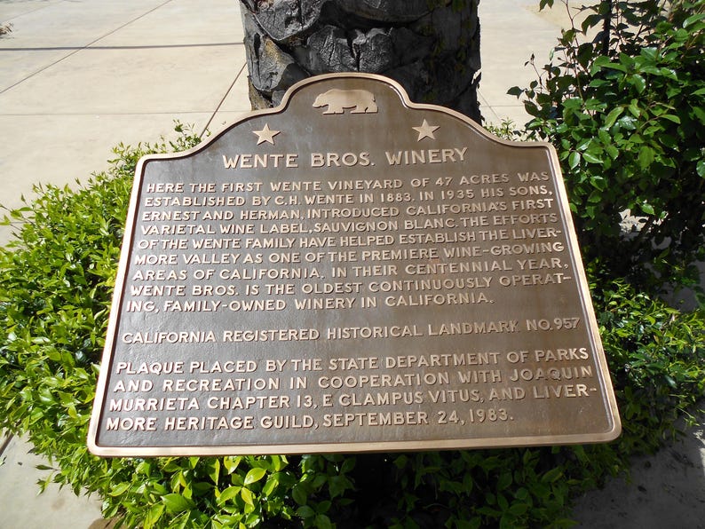 Puede incluir: Una placa hist&oacute;rica de metal color bronce titulada Wente Bros. Winery. La placa presenta un emblema de oso, dos estrellas y texto en relieve que detalla la historia del vi&ntilde;edo establecido en 1883, designado como California Landmark No. 957.