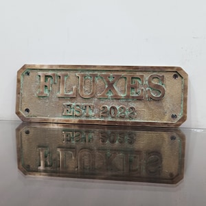 May include: A rectangular brass sign with rounded corners and a green patina. The sign reads "FLUXES EST 2022".