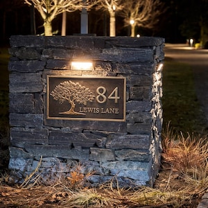 May include: A rectangular bronze address plaque mounted on a dark gray stacked stone pillar. The plaque features a raised relief of a tree, the number 84, and the text LEWIS LANE. The sign is illuminated by a small light fixture above it.