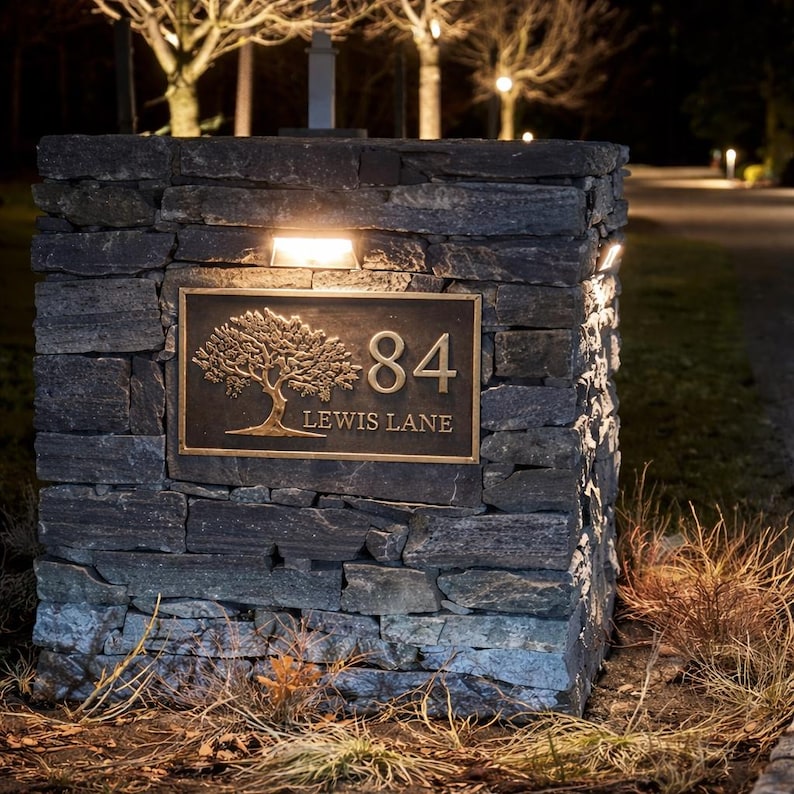 May include: A rectangular bronze address plaque mounted on a dark gray stacked stone pillar. The plaque features a raised relief of a tree, the number 84, and the text LEWIS LANE. The sign is illuminated by a small light fixture above it.
