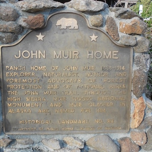 Puede incluir: Una placa hist&oacute;rica de bronce titulada JOHN MUIR HOME, con un emblema de oso y dos estrellas en la parte superior. El texto detalla la vida de John Muir, su defensa de la protecci&oacute;n forestal y los lugares nombrados en su honor, Landmark No. 312.