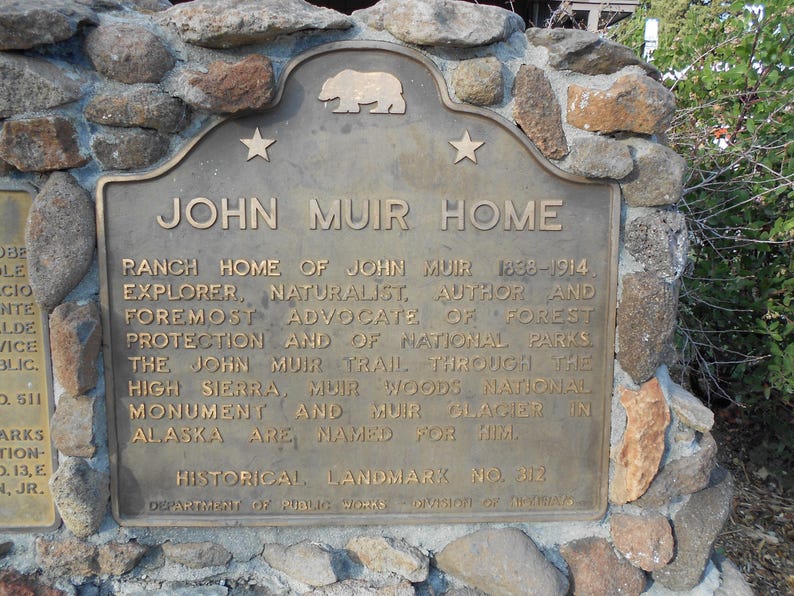 Puede incluir: Una placa hist&oacute;rica de bronce titulada JOHN MUIR HOME, con un emblema de oso y dos estrellas en la parte superior. El texto detalla la vida de John Muir, su defensa de la protecci&oacute;n forestal y los lugares nombrados en su honor, Landmark No. 312.