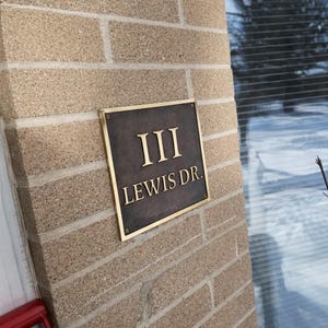 May include: A square metal address plaque with a dark, textured finish and a polished gold-toned border. The plaque features the raised gold-toned text "III" on the top line and "LEWIS DR." on the bottom line, secured with four corner screws.