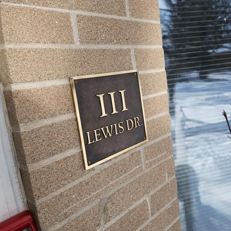 May include: A square metal address plaque with a dark, textured finish and a polished gold-toned border. The plaque features the raised gold-toned text "III" on the top line and "LEWIS DR." on the bottom line, secured with four corner screws.