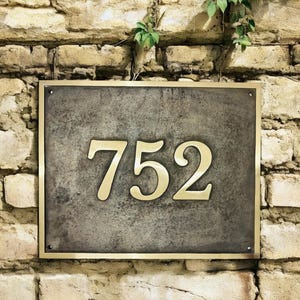 May include: A rectangular house number plaque featuring the digits 752 in a polished gold-tone finish. The numbers are mounted on a dark, weathered, textured gray metal plate, which is framed by a thin, matching polished gold-tone metal border.