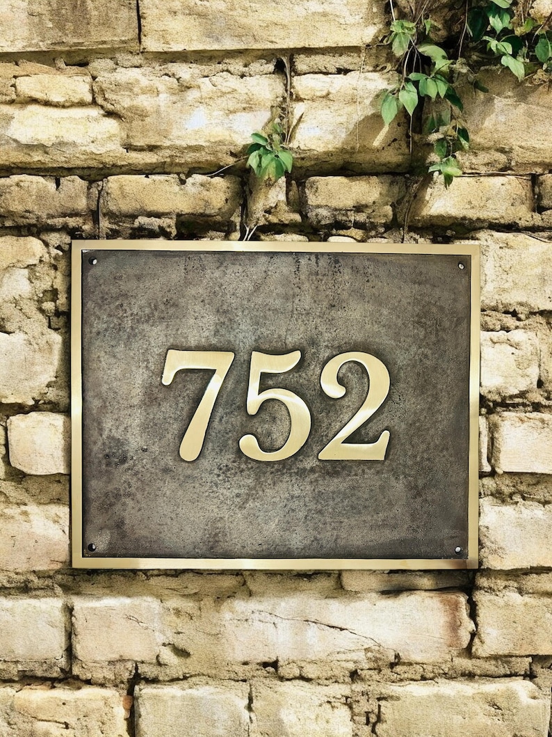 May include: A rectangular house number plaque featuring the digits 752 in a polished gold-tone finish. The numbers are mounted on a dark, weathered, textured gray metal plate, which is framed by a thin, matching polished gold-tone metal border.