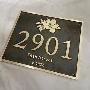 May include: A rectangular metal house plaque with a dark gray finish and a polished gold border. The plaque features a gold floral emblem above the house number 2901, followed by the text 34th Street and c. 1922 in gold lettering.
