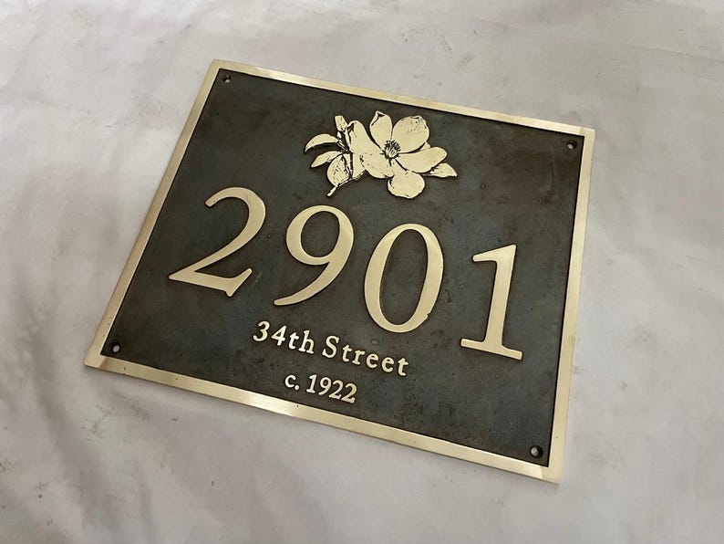 May include: A rectangular metal house plaque with a dark gray finish and a polished gold border. The plaque features a gold floral emblem above the house number 2901, followed by the text 34th Street and c. 1922 in gold lettering.