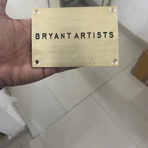 May include: A rectangular brass plaque with the text "BRYANT ARTISTS" in black lettering. The plaque has four drilled holes, one in each corner. The plaque is held in a hand.