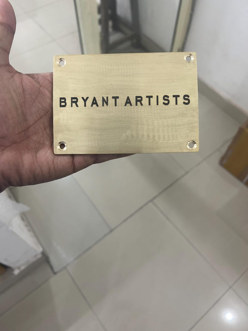 May include: A rectangular brass plaque with the text "BRYANT ARTISTS" in black lettering. The plaque has four drilled holes, one in each corner. The plaque is held in a hand.