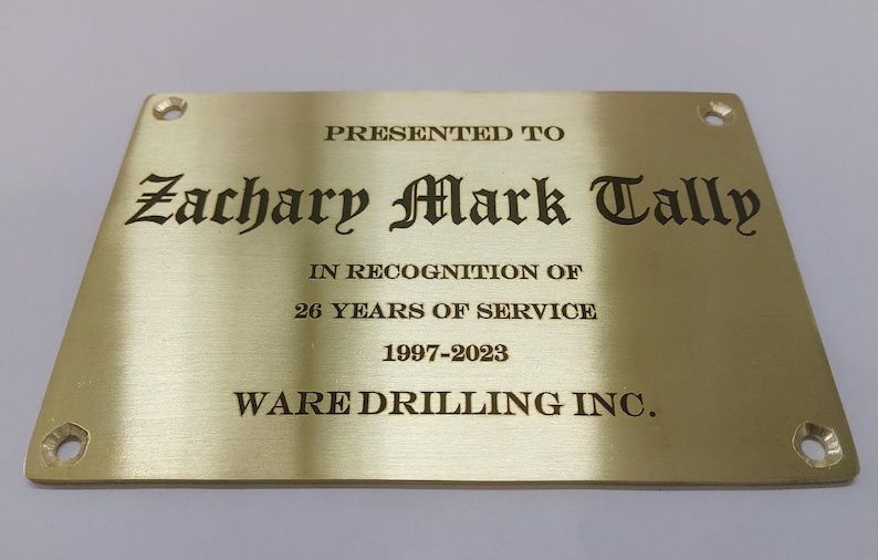 May include: A rectangular brass plaque with rounded corners and four mounting holes. The plaque features engraved text: "PRESENTED TO Zachary Mark Tally IN RECOGNITION OF 26 YEARS OF SERVICE 1997-2023 WARE DRILLING INC."