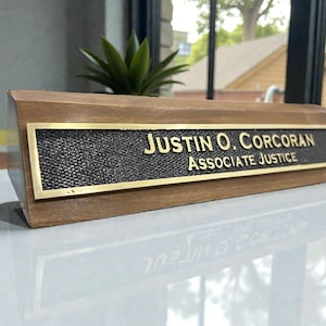 May include: A triangular wooden desk nameplate featuring a rectangular metal plaque with a textured black background and gold lettering. The text reads JUSTIN O. CORCORAN and ASSOCIATE JUSTICE in a clean, sans-serif font with a gold border.