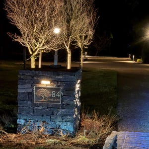 May include: A rectangular stone pillar features a dark metal plaque with the number 84 and the text LEWIS LANE, accompanied by an engraved tree illustration. The pillar is illuminated by a small light fixture mounted above the plaque at night.