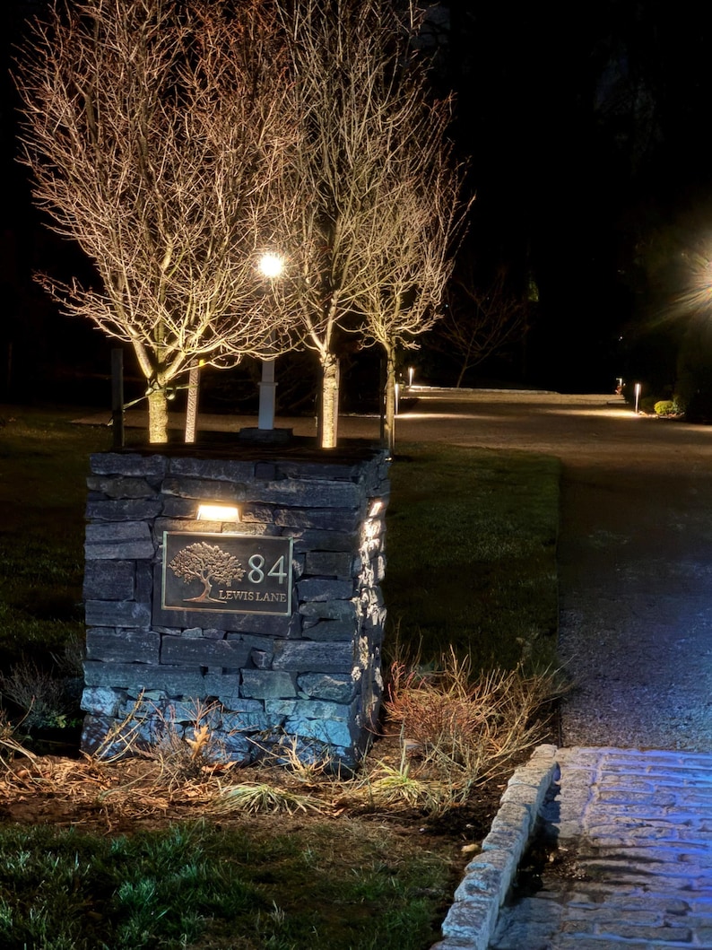 May include: A rectangular stone pillar features a dark metal plaque with the number 84 and the text LEWIS LANE, accompanied by an engraved tree illustration. The pillar is illuminated by a small light fixture mounted above the plaque at night.