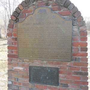 Puede incluir: Un monumento de ladrillo rojo con una placa de bronce titulada Mount Diablo Coal Field. La placa incluye un emblema de oso, dos estrellas y texto que detalla la historia del distrito minero, adem&aacute;s de una placa secundaria m&aacute;s peque&ntilde;a debajo.