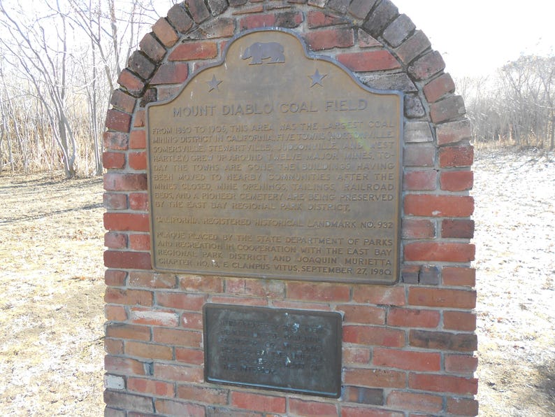 Puede incluir: Un monumento de ladrillo rojo con una placa de bronce titulada Mount Diablo Coal Field. La placa incluye un emblema de oso, dos estrellas y texto que detalla la historia del distrito minero, adem&aacute;s de una placa secundaria m&aacute;s peque&ntilde;a debajo.