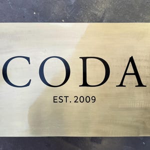 May include: A rectangular brass sign with the word "CODA" in bold black letters. Below the word is the text "EST. 2009" in a smaller, black font. The sign has a brushed finish and is mounted with four small screws.